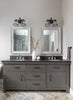 Bathroom vanity with two mirrors, lights, and decorative items on a neutral wall.