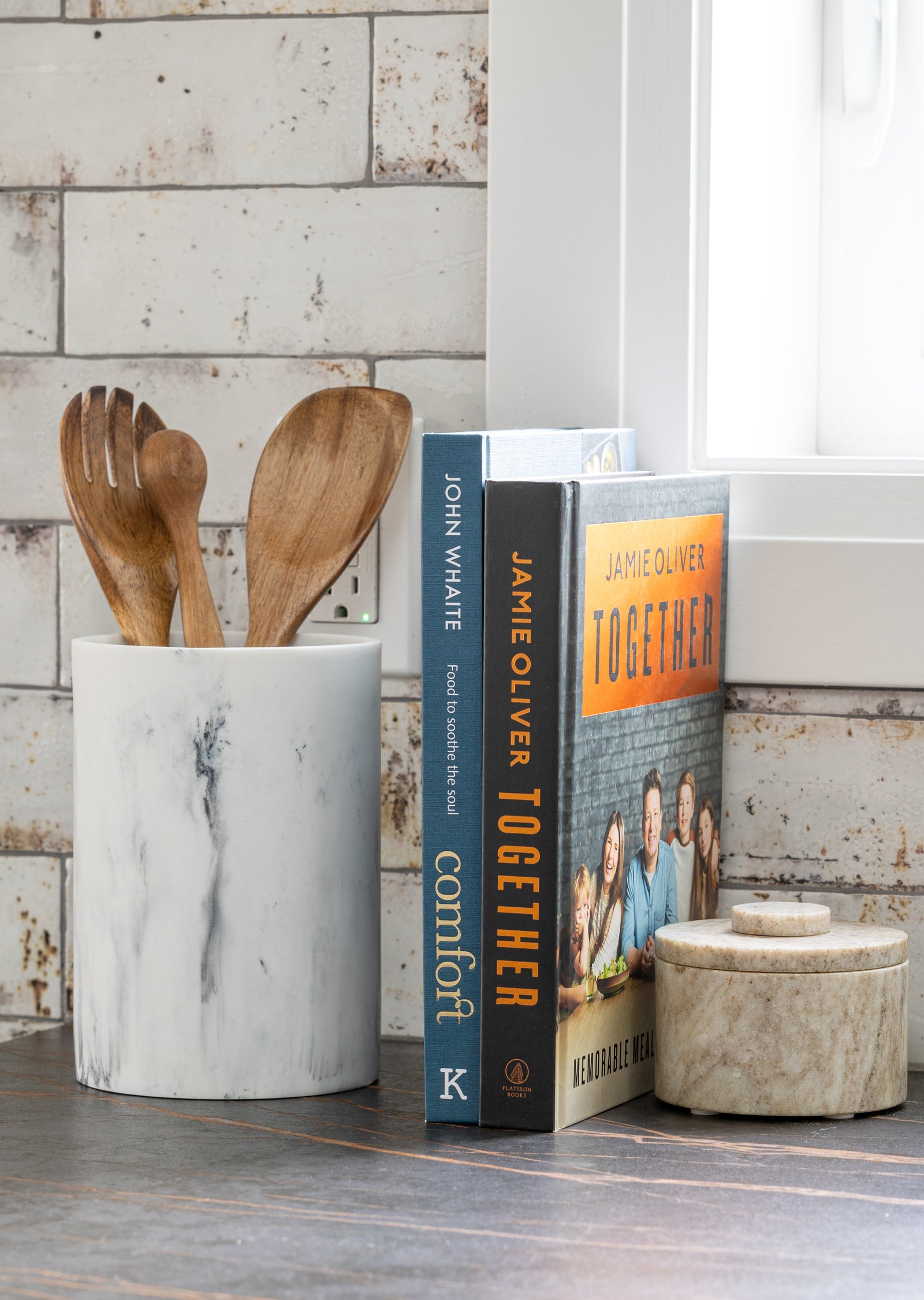 Marble utensil holder with wooden spoons next to cookbooks on a wooden surface.
