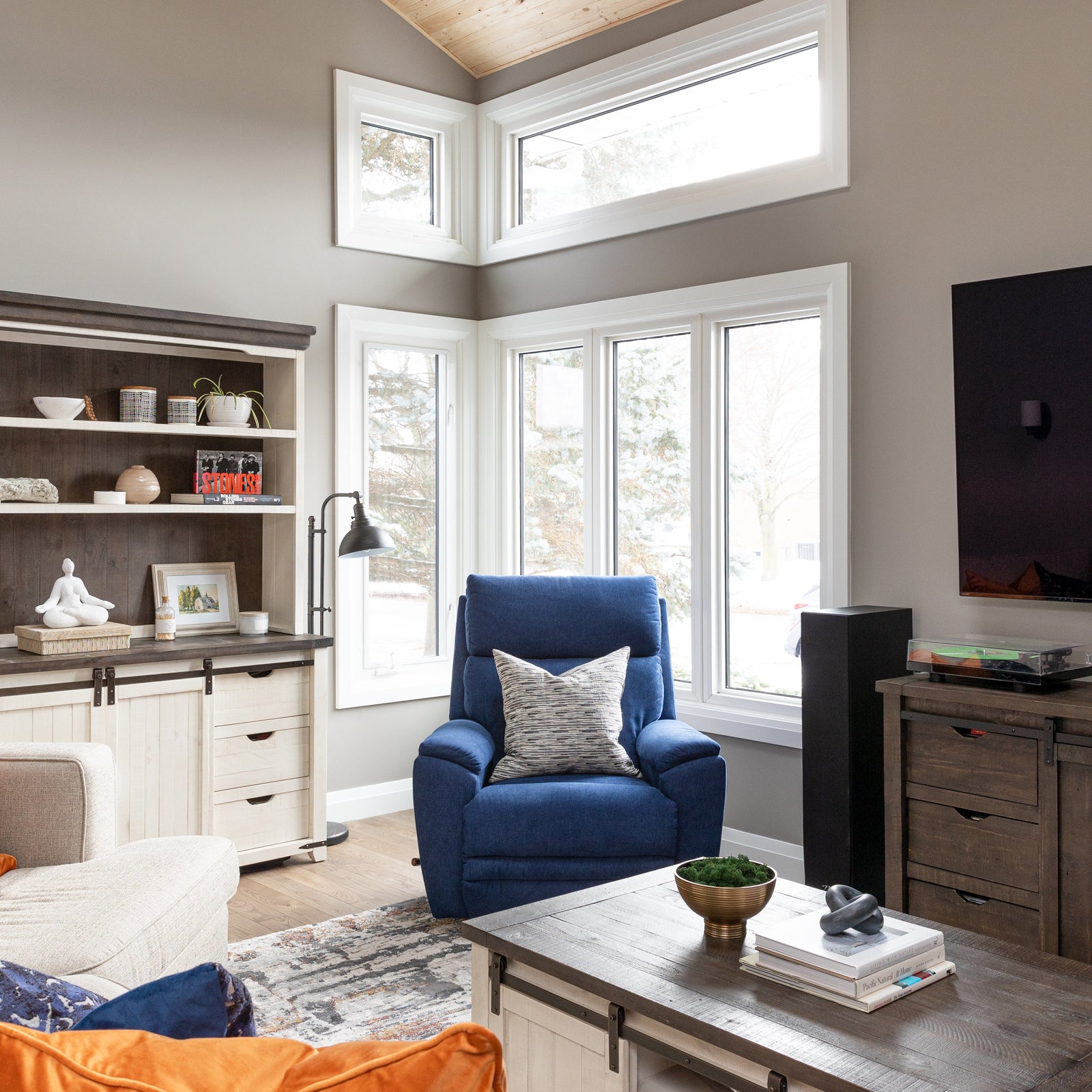 Modern living room with a blue armchair, orange pouf, and large windows.