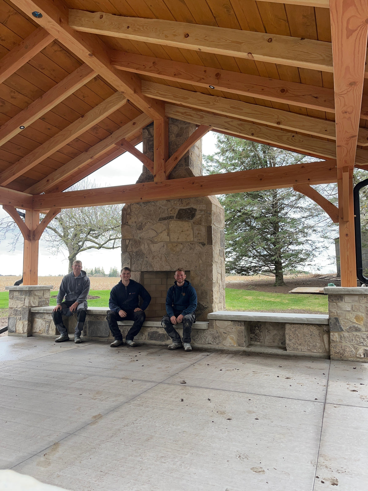 People sitting under a wooden pavilion with a stone wall and grassy area in the background.