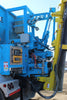 Blue truck-mounted hydraulic equipment system with control panel, hoses, and mechanical arm attached to yellow industrial vehicle on job site