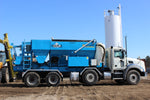 Truck-mounted stone slinger with large blue aggregate hopper and conveyor system parked on construction site with vertical storage silo in background