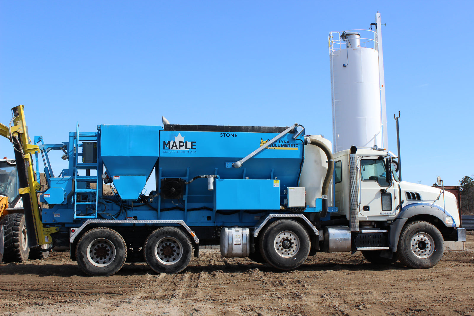 Truck-mounted stone slinger with large blue aggregate hopper and conveyor system parked on construction site with vertical storage silo in background