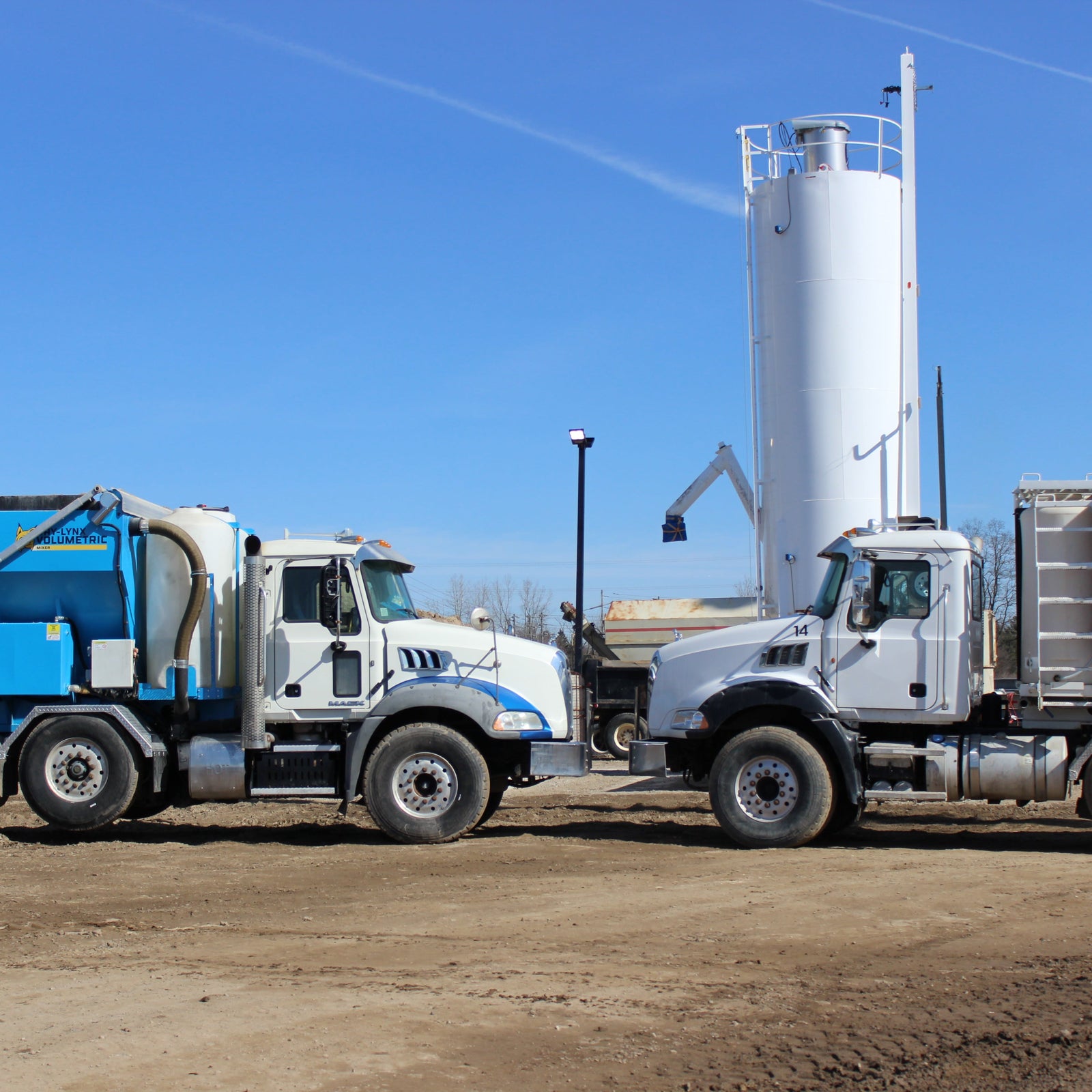 Two volumetric concrete trucks parked on a construction site with a large white silo in the background under a clear blue sky.