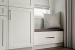 Window seat with a cushion in a room with white cabinets and curtains.