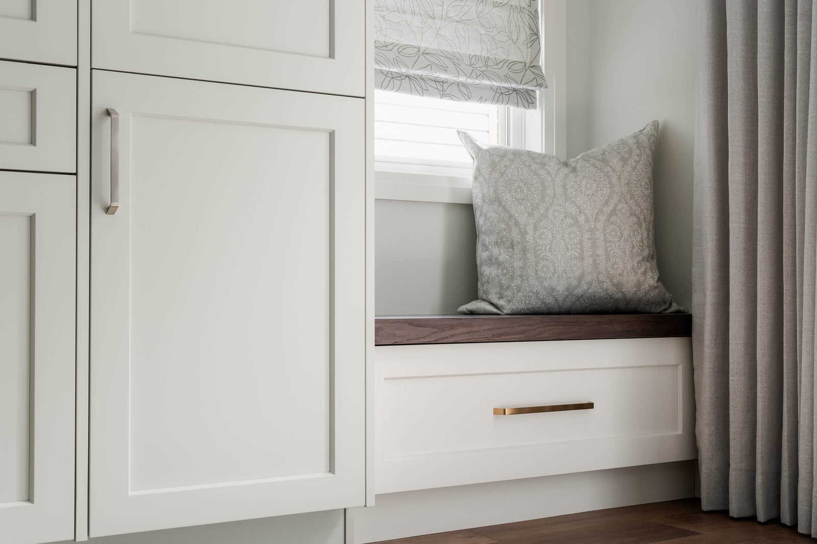 Window seat with a cushion in a room with white cabinets and curtains.