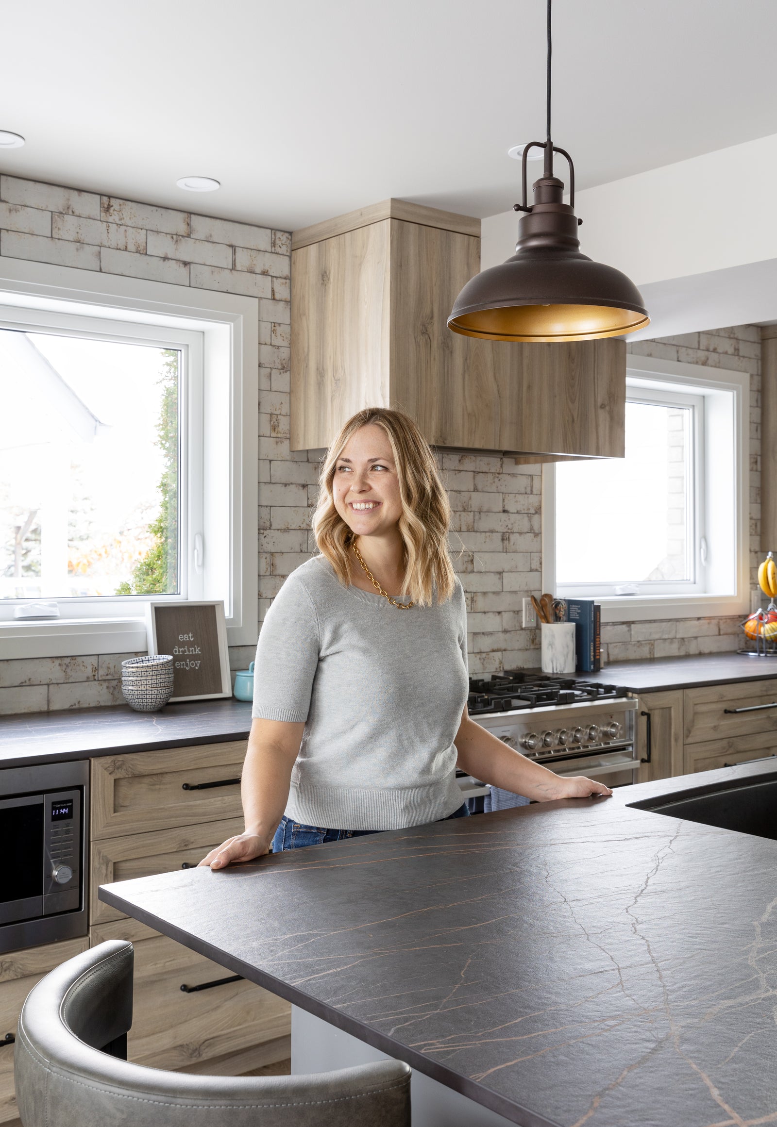 Woman standing in a modern kitchen with wooden cabinets and a pendant light.